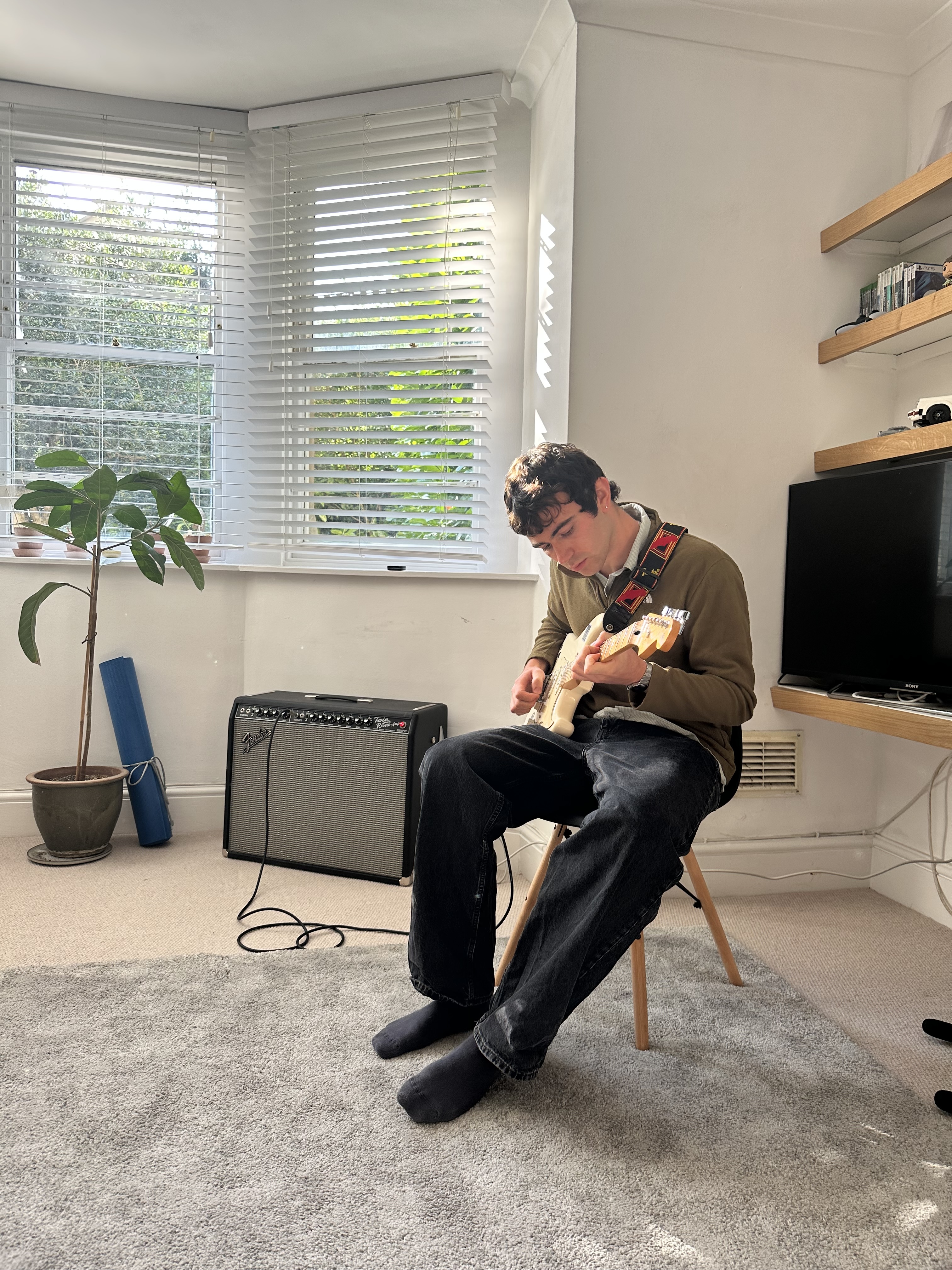 Man sitting in a white room, playing electric guitar on a stool.